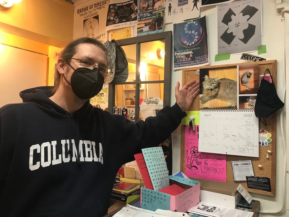 Volume 2 collective member Ryan Licwinko poses behind the back desk next to the customized 12-month calendar of his cat Max