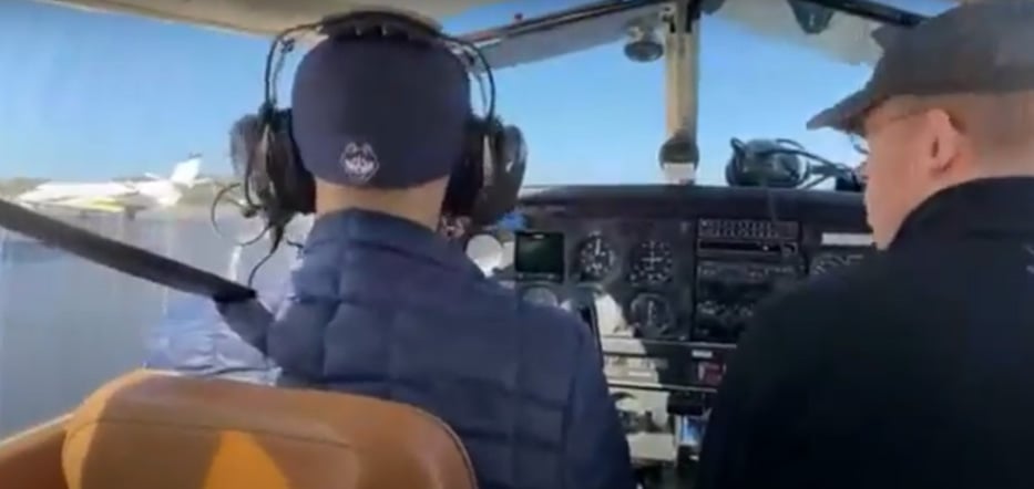 Two men sit in the cockpit of a plane. The man on the left is dressed in a blue hat and jacket. The man on the right is also dressed in a blue hat and sweater.