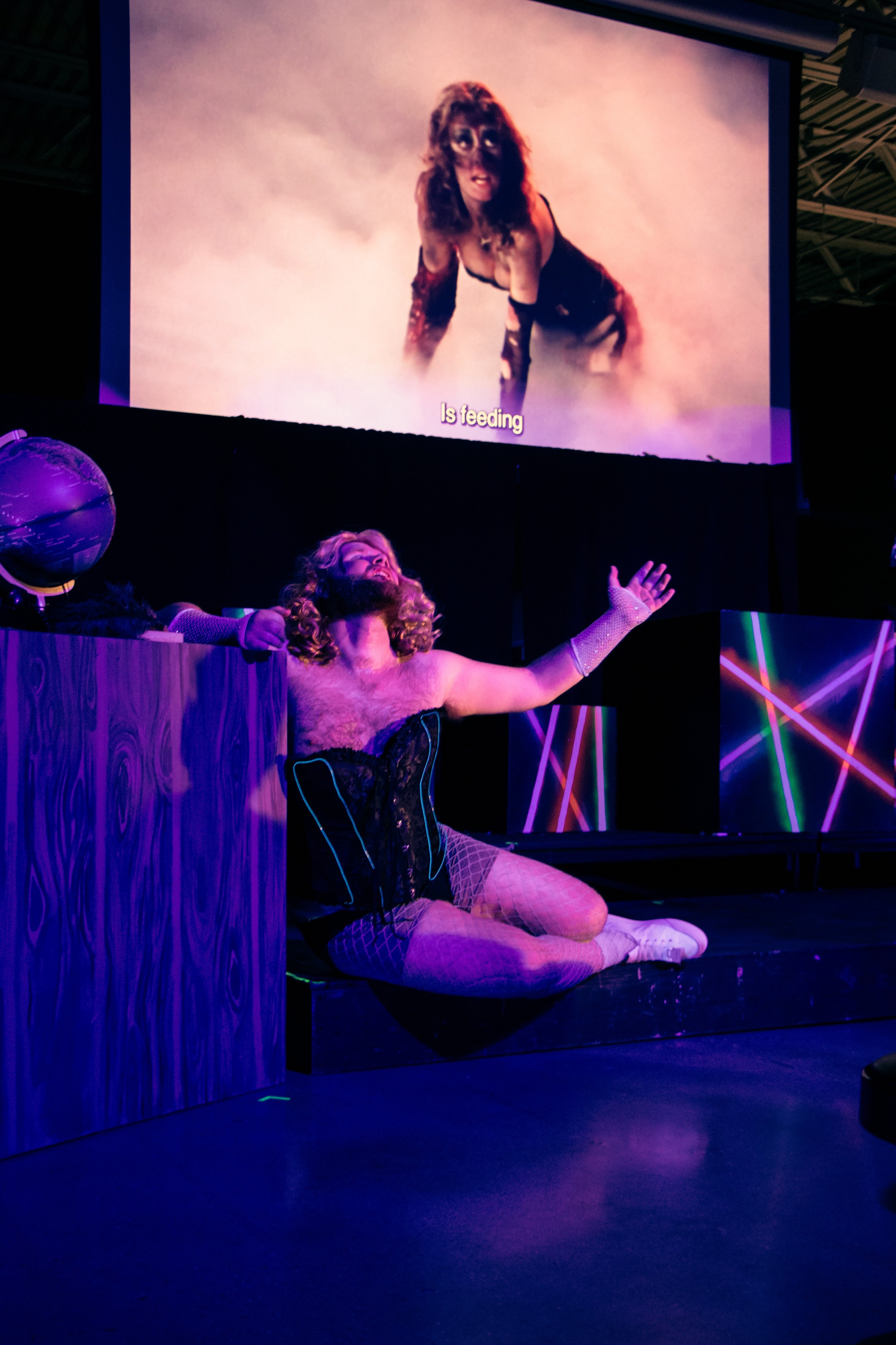 A performer wearing a corset and a wig sings on stage under a projection of a film where an actor wearing the same costume sings as captions read "Is feeding" || Jacob Marks as Janet Weiss (Picture credit: Tony DeZinno)